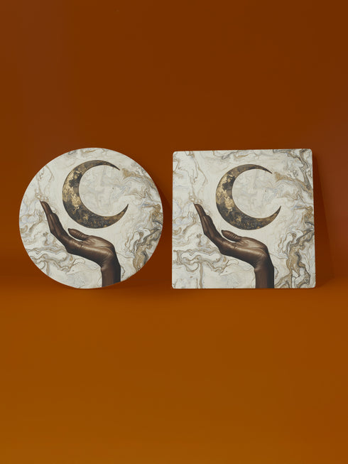 A ceramic coaster featuring a bronze-colored hand reaching toward an antiqued gold crescent moon against a swirling ivory and gold marble background, available in round and square shapes with cork backing.
